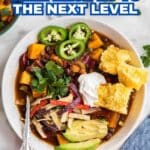 A bowl of chili with beans, sweet potatoes, tortilla strips, avocado, jalapeño slices, sour cream, cilantro, and cornbread, shown on a table with chili and cornbread on the side.