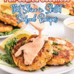 Two pan-seared seafood patties on a white plate, topped with creamy sauce, served with arugula and lemon wedges. Text overlay highlights skillet seafood recipes packed with protein.