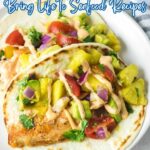 Two fish tacos topped with pineapple salsa, diced red onions, cherry tomatoes, fresh herbs, and a drizzle of creamy sauce bring a citrus twist to classic seafood recipes on a white plate.
