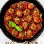 A cast iron skillet filled with meatballs in red tomato sauce, garnished with fresh basil leaves and chopped herbs.