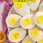 Plate of oval-shaped cookies decorated to look like deviled eggs, with white icing and yellow frosting centers, on a pink cloth with lemon slices and purple flowers nearby.
