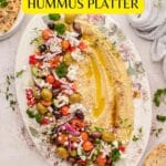 Oval platter with hummus topped with olives, cherry tomatoes, feta, herbs, and spices, surrounded by pita bread and garnished with parsley.