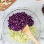 A glass bowl with shredded purple and green cabbage, mixed with a wooden spoon. Surrounding bowls contain shredded carrots, apples, and beets.