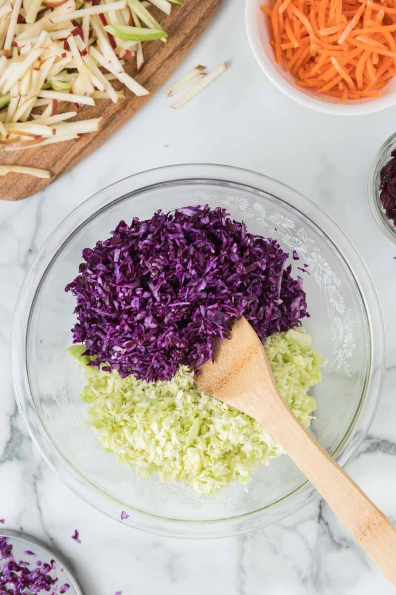 A glass bowl with shredded purple and green cabbage, mixed with a wooden spoon. Surrounding bowls contain shredded carrots, apples, and beets.