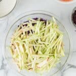 A glass bowl of julienned green apples on a marble surface, surrounded by bowls of shredded carrots, seeds, dressing, and chopped green onions.