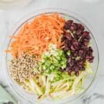 A glass bowl containing shredded carrots, dried cranberries, chopped green onions, sunflower seeds, and julienned green apple on a marble surface.