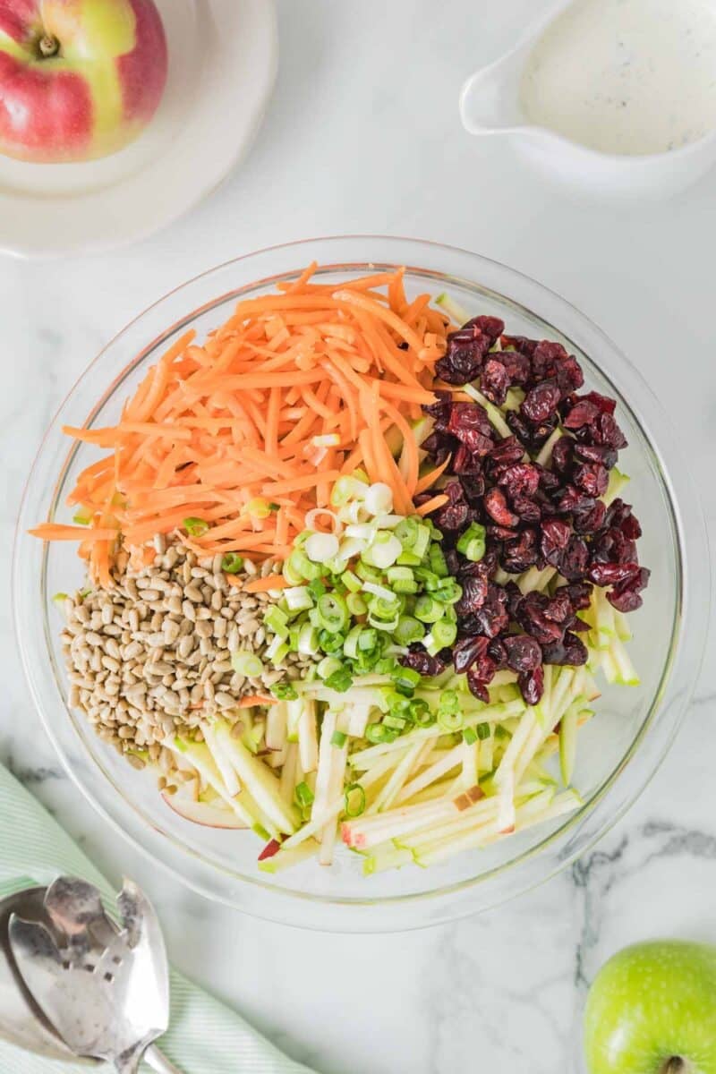 A glass bowl containing shredded carrots, dried cranberries, chopped green onions, sunflower seeds, and julienned green apple on a marble surface.