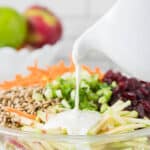 A bowl of salad with shredded carrots, green onions, sunflower seeds, dried cranberries, and apple slices being topped with creamy dressing from a white pitcher.