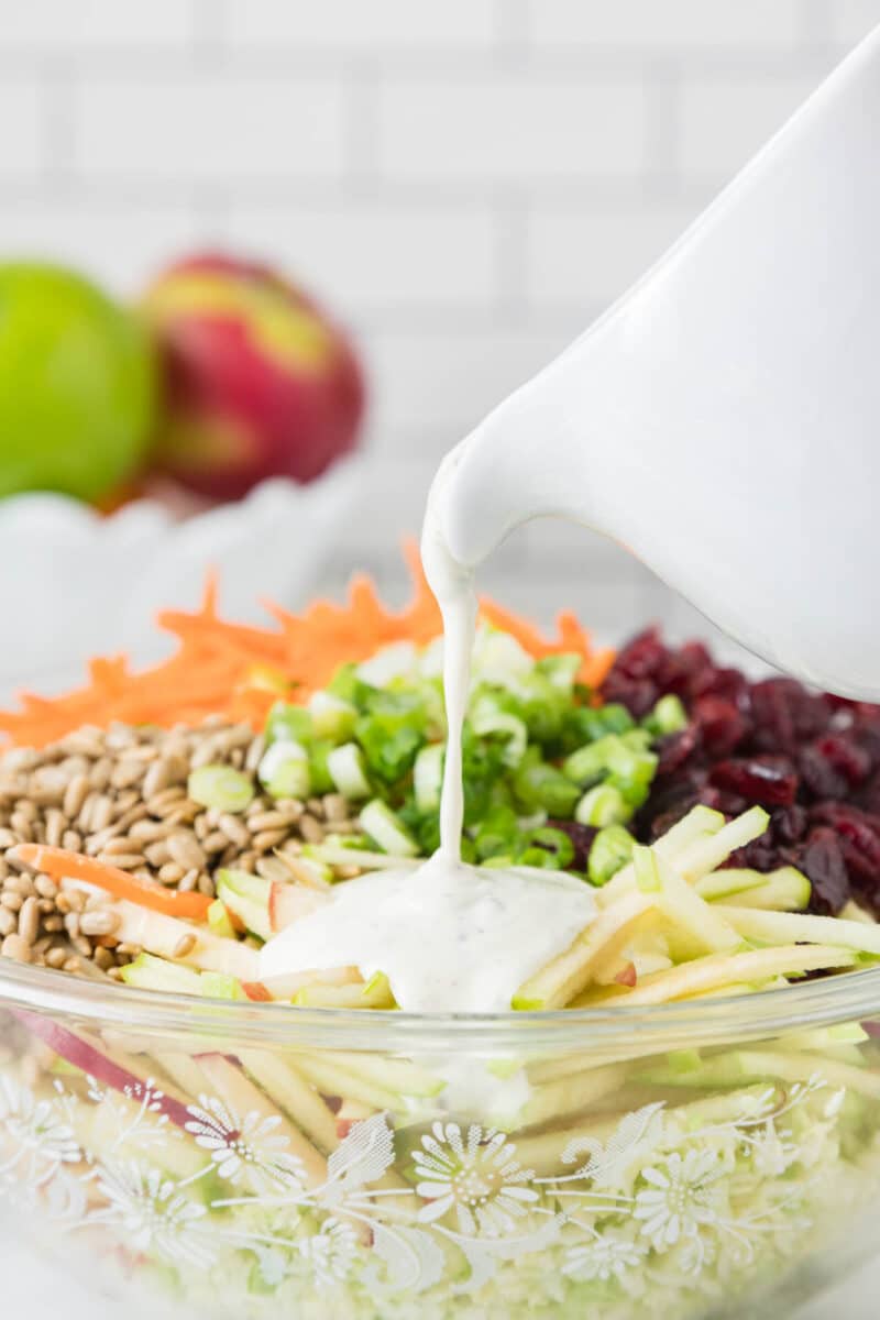 A bowl of salad with shredded carrots, green onions, sunflower seeds, dried cranberries, and apple slices being topped with creamy dressing from a white pitcher.