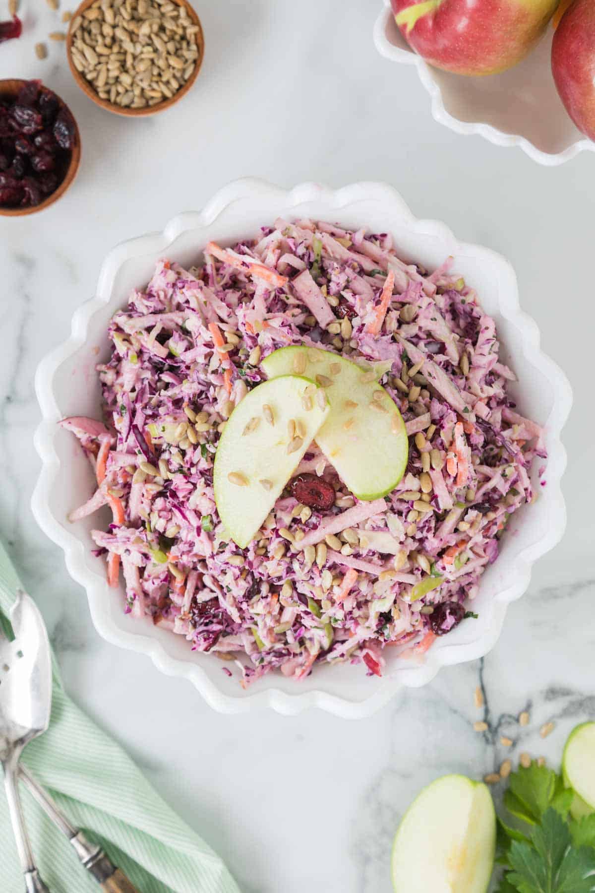 A bowl of apple coleslaw made with purple cabbage, carrots, and seeds, garnished with apple slices, on a marble surface with apples, seeds, and dried cranberries beside it.