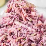 A spoonful of apple coleslaw with shredded red cabbage, carrots, and dressing is held above a bowl filled with more coleslaw.