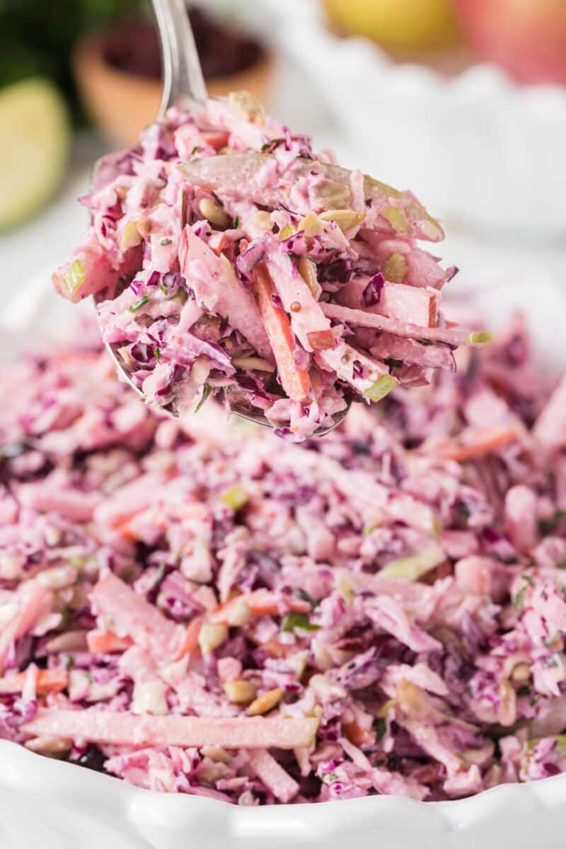 A spoonful of apple coleslaw with shredded red cabbage, carrots, and dressing is held above a bowl filled with more coleslaw.