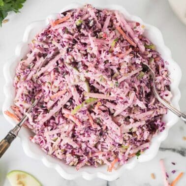 A bowl of coleslaw with shredded red cabbage, carrots, and green apple, mixed with a creamy dressing. Two serving utensils are in the bowl, and apples are sliced nearby.