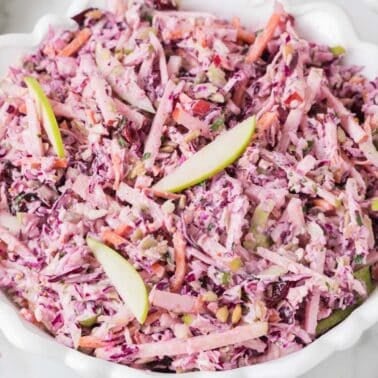 A bowl of coleslaw with shredded red cabbage, apples, carrots, and a creamy dressing, garnished with thin apple slices.