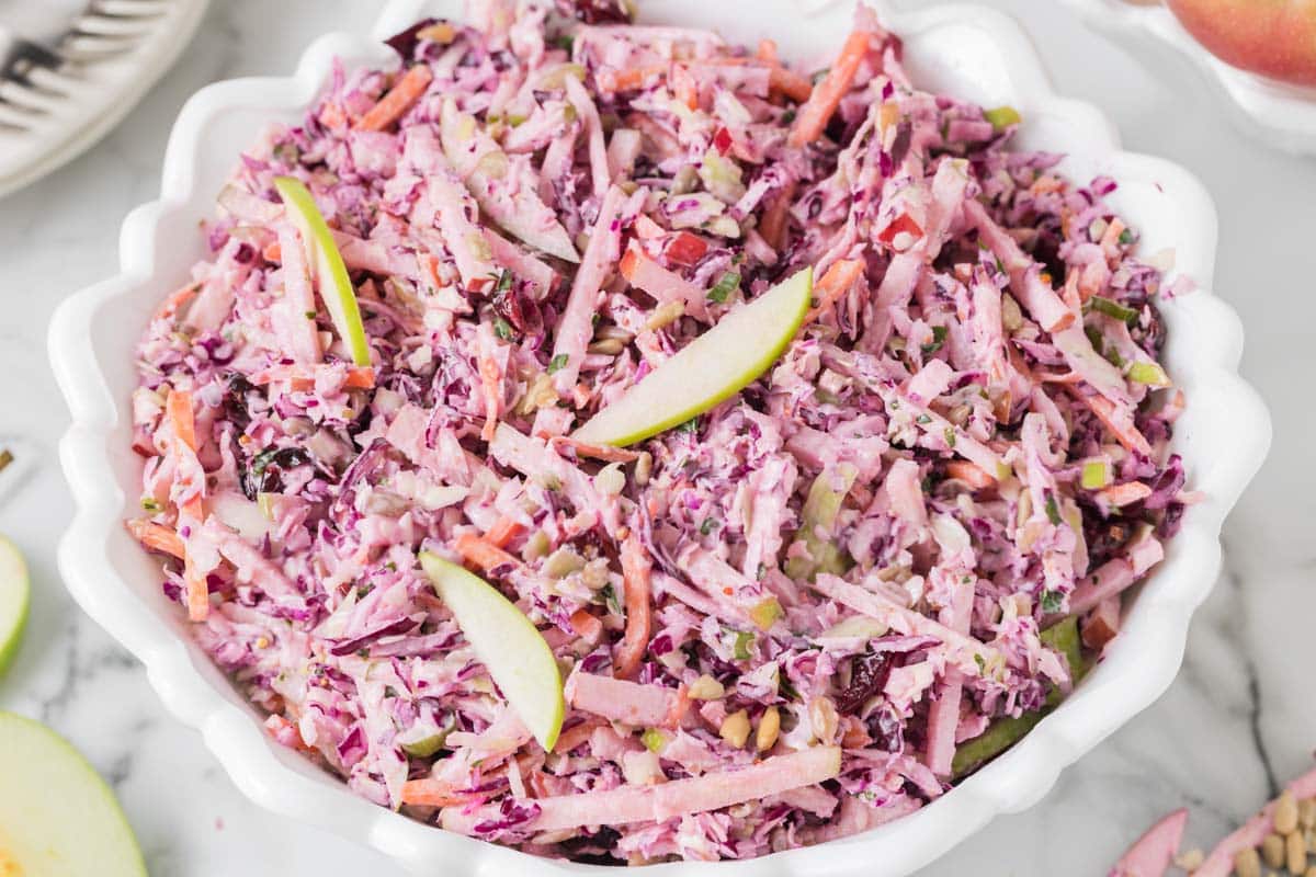 A bowl of coleslaw with shredded red cabbage, apples, carrots, and a creamy dressing, garnished with thin apple slices.