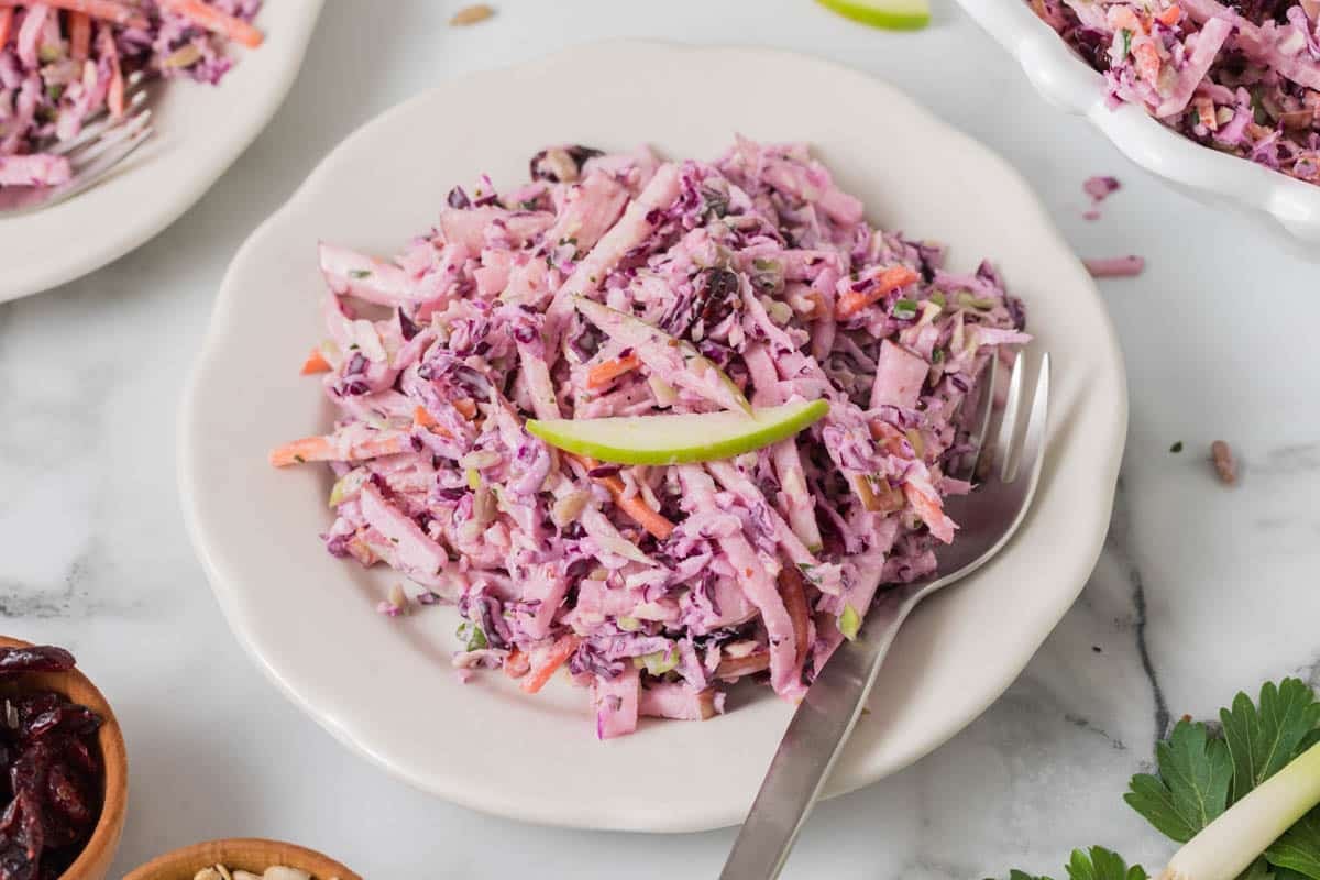 A plate of creamy coleslaw with shredded red cabbage, carrots, and green apple, garnished with a thin apple slice, with a fork on the side.