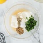 A glass bowl with mayonnaise, chopped herbs, whole grain mustard, salt, pepper, and lemon juice on a marble countertop, surrounded by a whisk, a strainer, and a squeezed lemon.