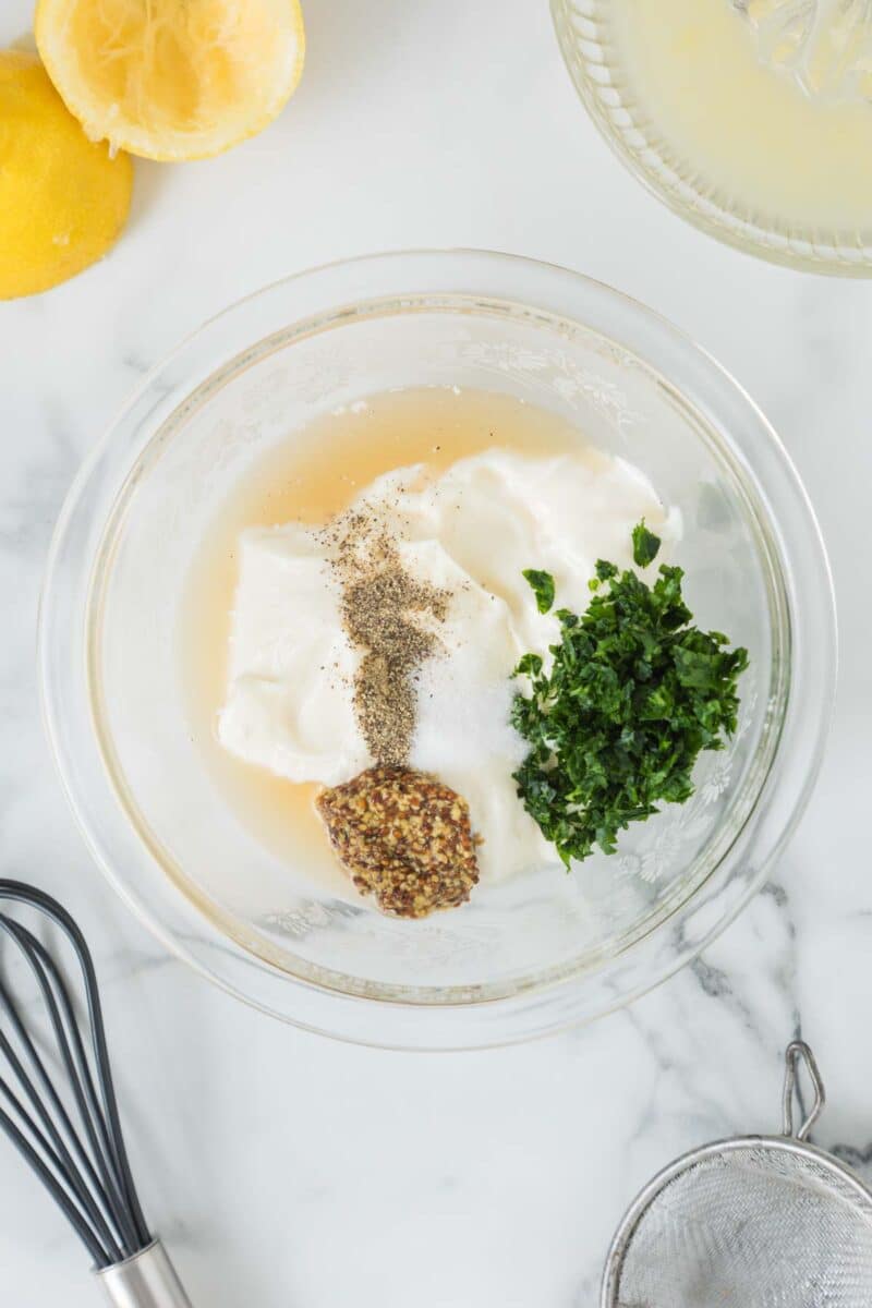 A glass bowl with mayonnaise, chopped herbs, whole grain mustard, salt, pepper, and lemon juice on a marble countertop, surrounded by a whisk, a strainer, and a squeezed lemon.