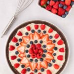 A chocolate dessert pizza topped with cream, strawberries, raspberries, and blueberries sits next to a square bowl of mixed berries and a metal whisk on a white surface.