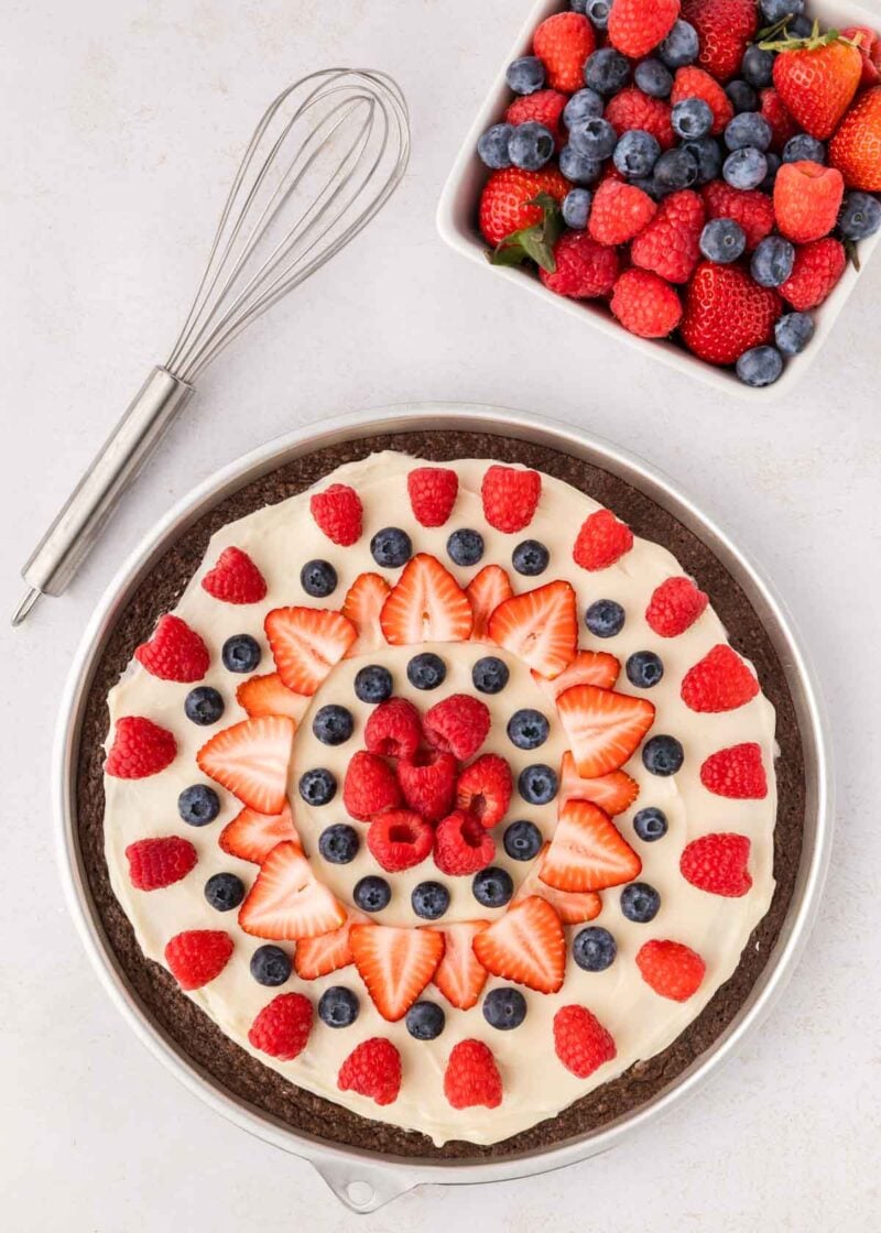 A chocolate dessert pizza topped with cream, strawberries, raspberries, and blueberries sits next to a square bowl of mixed berries and a metal whisk on a white surface.