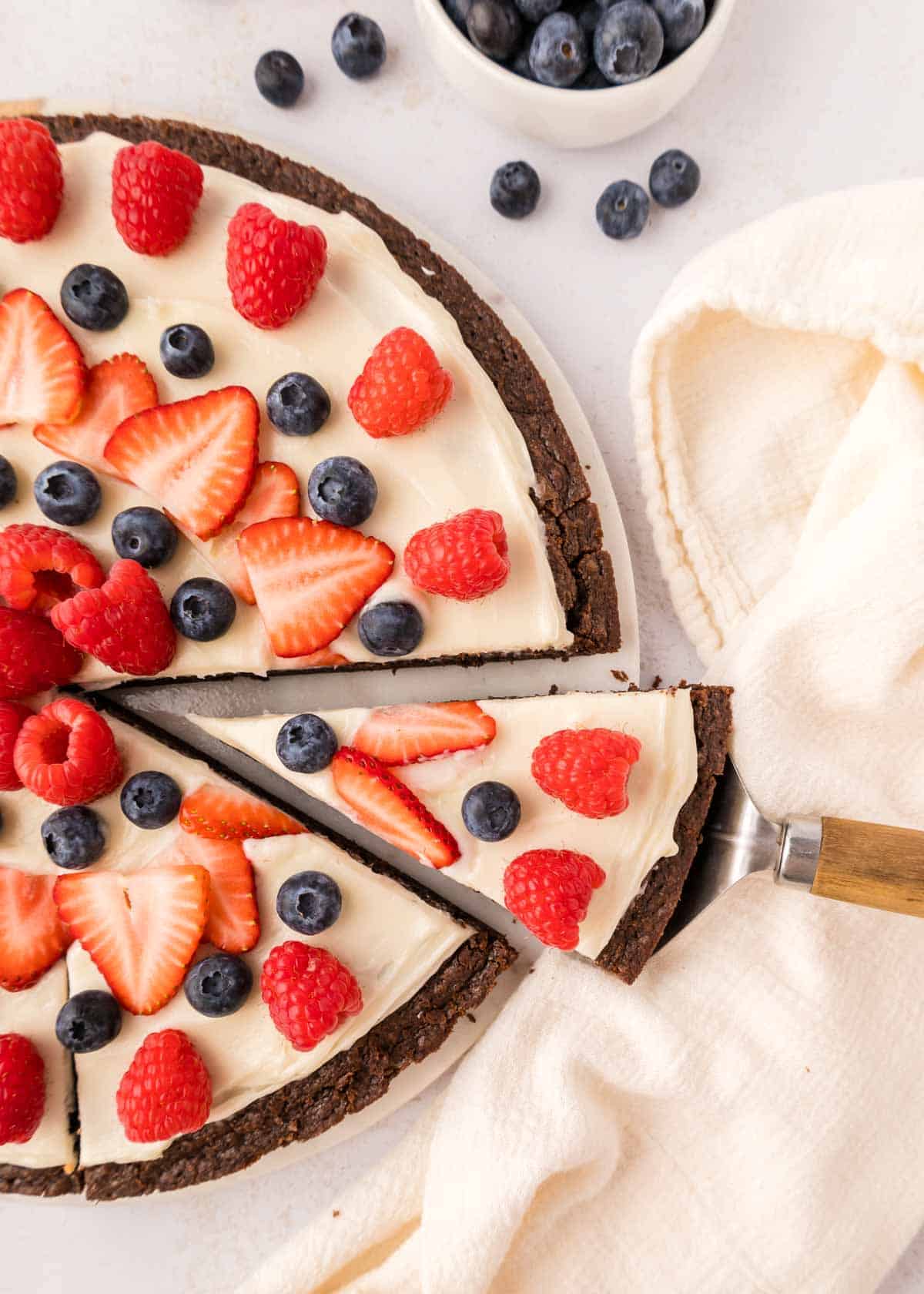 A chocolate dessert pizza topped with cream, fresh strawberries, raspberries, and blueberries is being sliced with a pie server next to a white cloth.