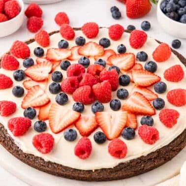 A large round dessert with a chocolate base, white frosting, and topped with fresh strawberries, raspberries, and blueberries. Bowls of berries are in the background.