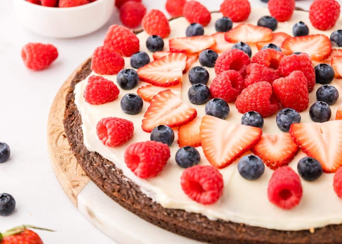 A dessert pizza with a chocolate crust, topped with white frosting, sliced strawberries, blueberries, and raspberries, displayed on a wooden board.