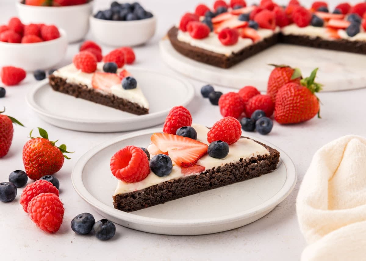 A slice of brownie topped with cream and fresh berries is served on a plate, with more slices, berries, and small bowls of fruit in the background.