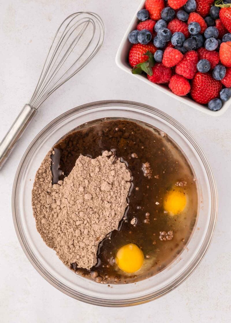 A glass bowl with brownie mix, two eggs, and liquid ingredients beside a whisk and a bowl of assorted fresh berries.