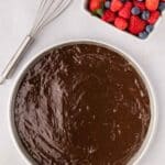 A mixing bowl filled with chocolate cake batter sits next to a whisk and a dish of assorted fresh berries, including strawberries, blueberries, and raspberries.
