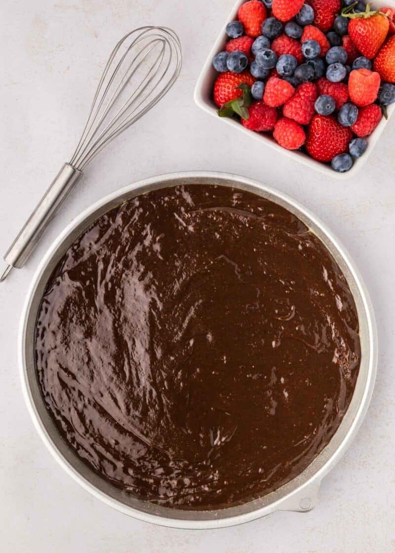 A mixing bowl filled with chocolate cake batter sits next to a whisk and a dish of assorted fresh berries, including strawberries, blueberries, and raspberries.