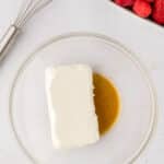 A glass bowl containing a block of cream cheese and a pool of vanilla extract, next to a whisk and a dish of assorted berries.