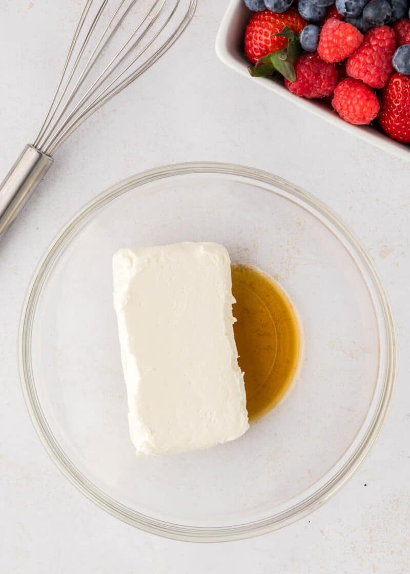 A glass bowl containing a block of cream cheese and a pool of vanilla extract, next to a whisk and a dish of assorted berries.