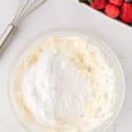 A glass bowl with creamed mixture and powdered sugar, a whisk beside it, and a bowl of fresh berries in the background.