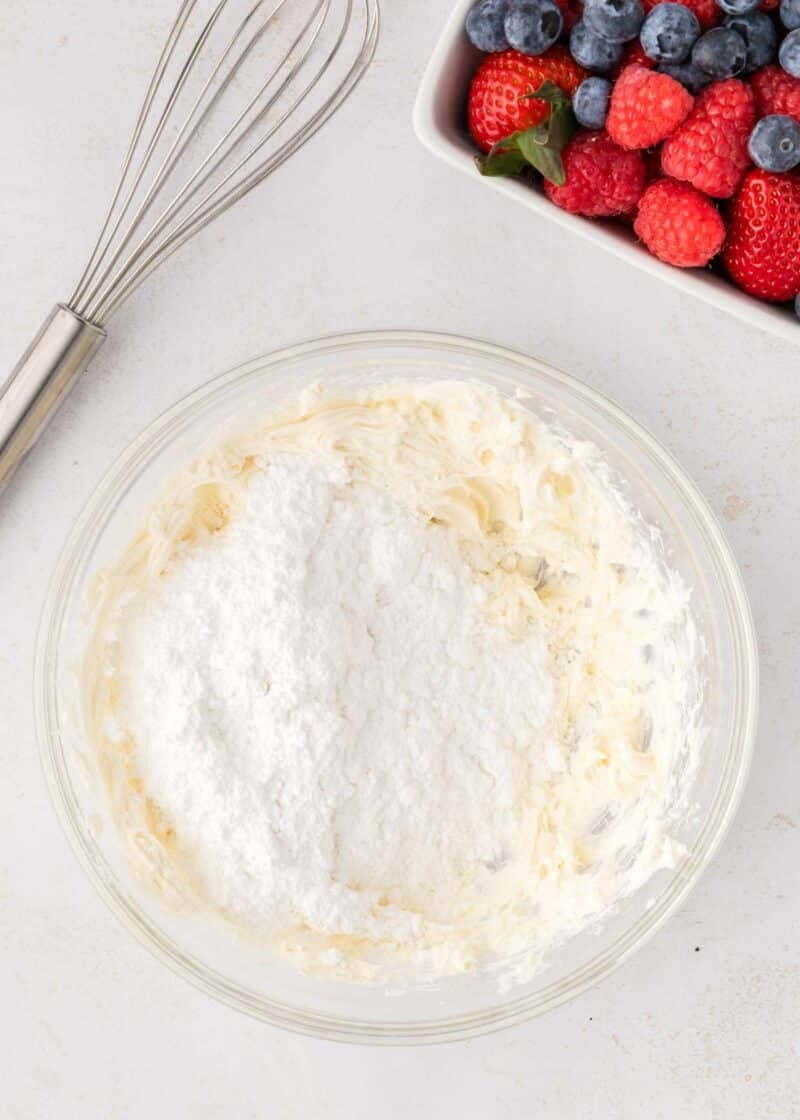 A glass bowl with creamed mixture and powdered sugar, a whisk beside it, and a bowl of fresh berries in the background.