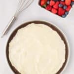 A chocolate dessert pizza with a layer of white frosting in a round pan, next to a metal whisk and a square bowl of assorted fresh berries.