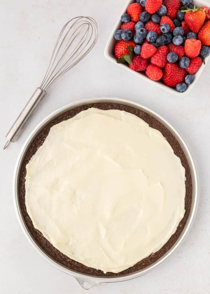 A chocolate dessert pizza with a layer of white frosting in a round pan, next to a metal whisk and a square bowl of assorted fresh berries.