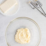 A glass bowl with softened cream cheese, a plate with a cream cheese block, and two metal beaters on a white countertop.