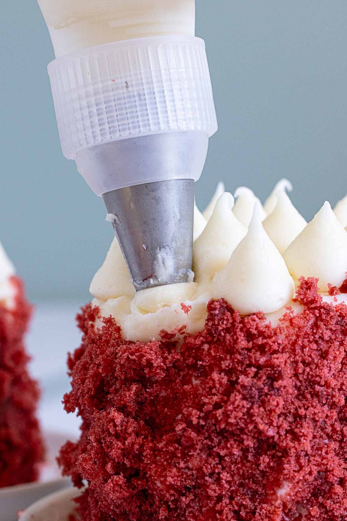 A piping bag is being used to decorate the top of a red velvet cake with dollops of white frosting.