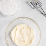 A glass bowl with creamed butter and sugar, a bowl of powdered sugar, and two metal beaters on a white surface.