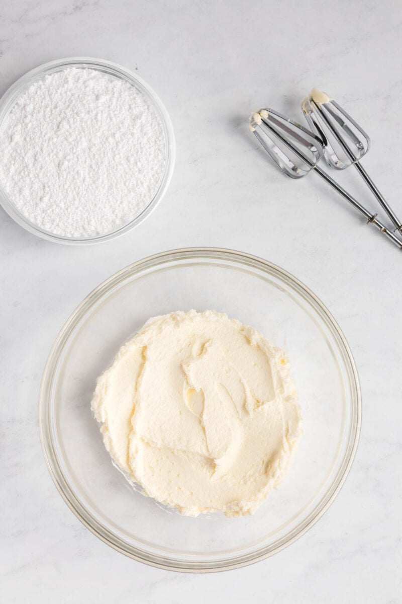 A glass bowl with creamed butter and sugar, a bowl of powdered sugar, and two metal beaters on a white surface.