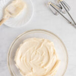 A glass bowl of creamy frosting sits on a white surface next to a plate with a spatula and electric mixer beaters.