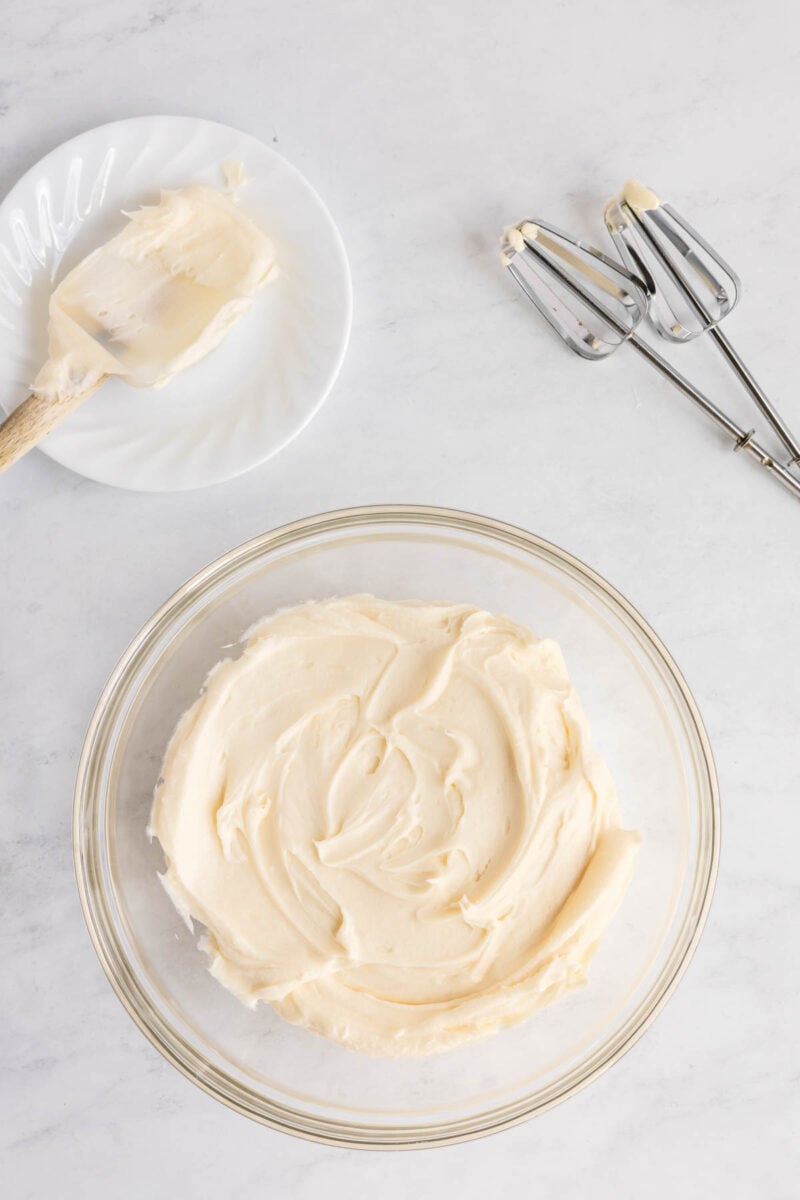 A glass bowl of creamy frosting sits on a white surface next to a plate with a spatula and electric mixer beaters.