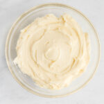 A glass bowl filled with smooth, creamy mashed potatoes on a light marble surface.