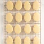 Sixteen plain, egg-shaped sugar cookies are arranged in a 4-by-4 grid on a cooling rack over a white marble surface.
