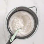A fine mesh strainer with powdered sugar is placed over a glass bowl, with a spatula resting inside the strainer on a white marble surface.