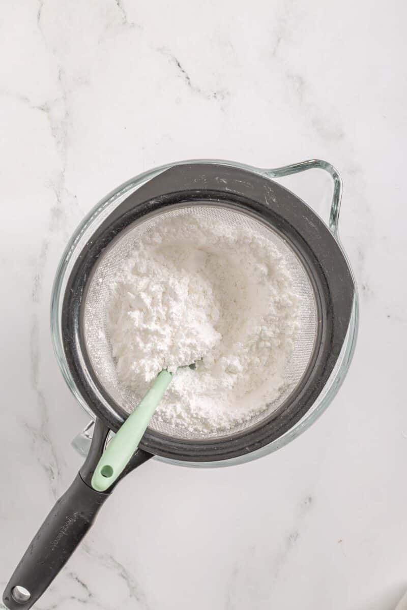 A fine mesh strainer with powdered sugar is placed over a glass bowl, with a spatula resting inside the strainer on a white marble surface.