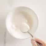 A gloved hand holds a spoon over a white bowl filled with a thick, white liquid on a marble surface.