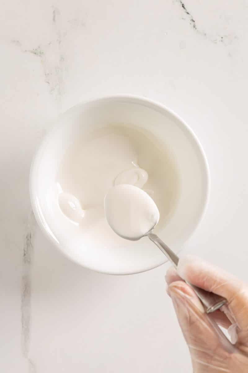 A gloved hand holds a spoon over a white bowl filled with a thick, white liquid on a marble surface.