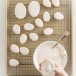 A gloved hand dips an oval cookie into a bowl of white icing, with iced oval cookies drying on a cooling rack in the background.